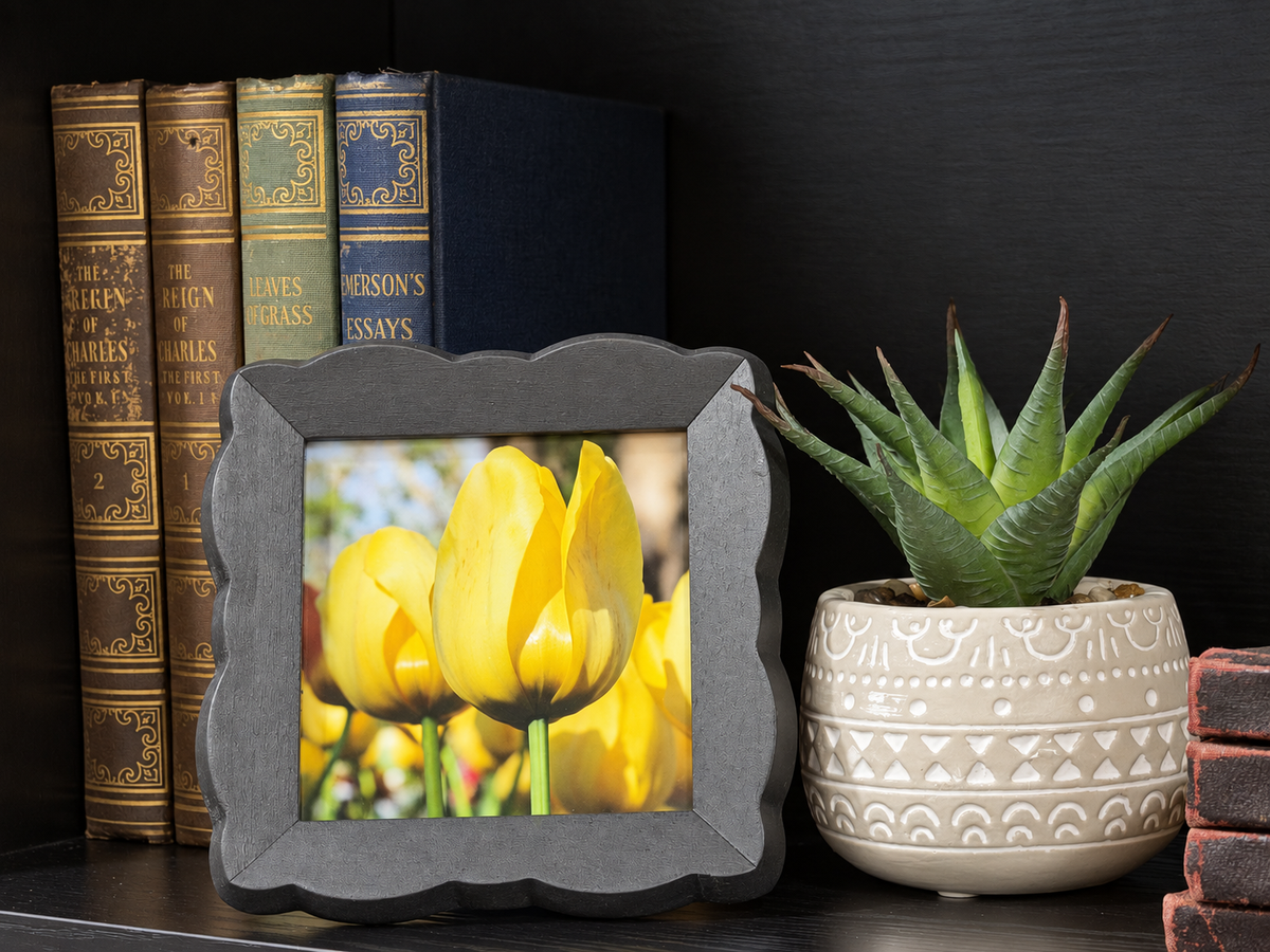 A scalloped dark gray picture frame displaying a photo of yellow tulips, styled on a dark wooden shelf next to a potted succulent and antique books.