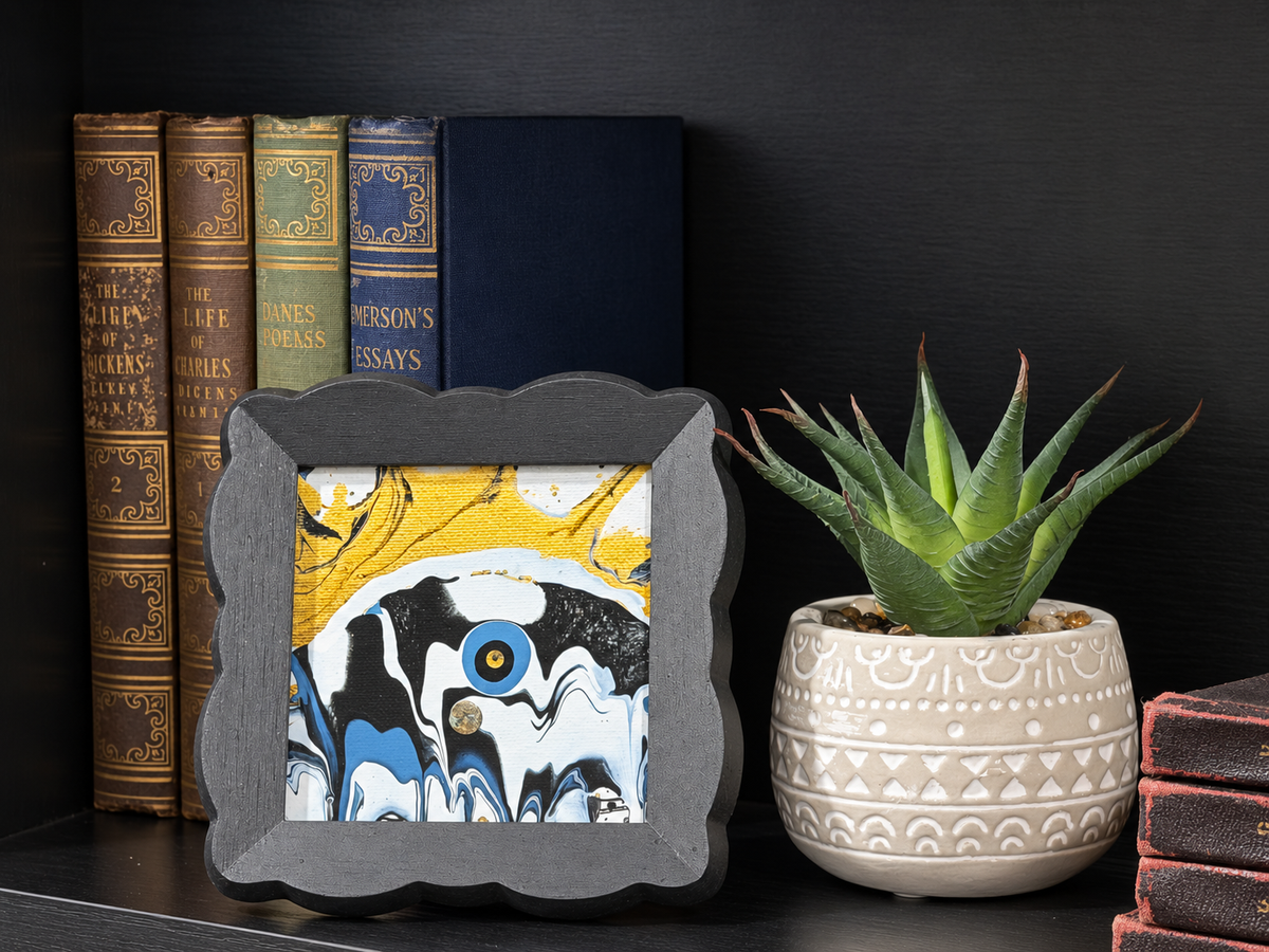 A square framed abstract art print with a marbled pattern of yellow, black, white, and blue, styled on a dark shelf next to a potted succulent and books.