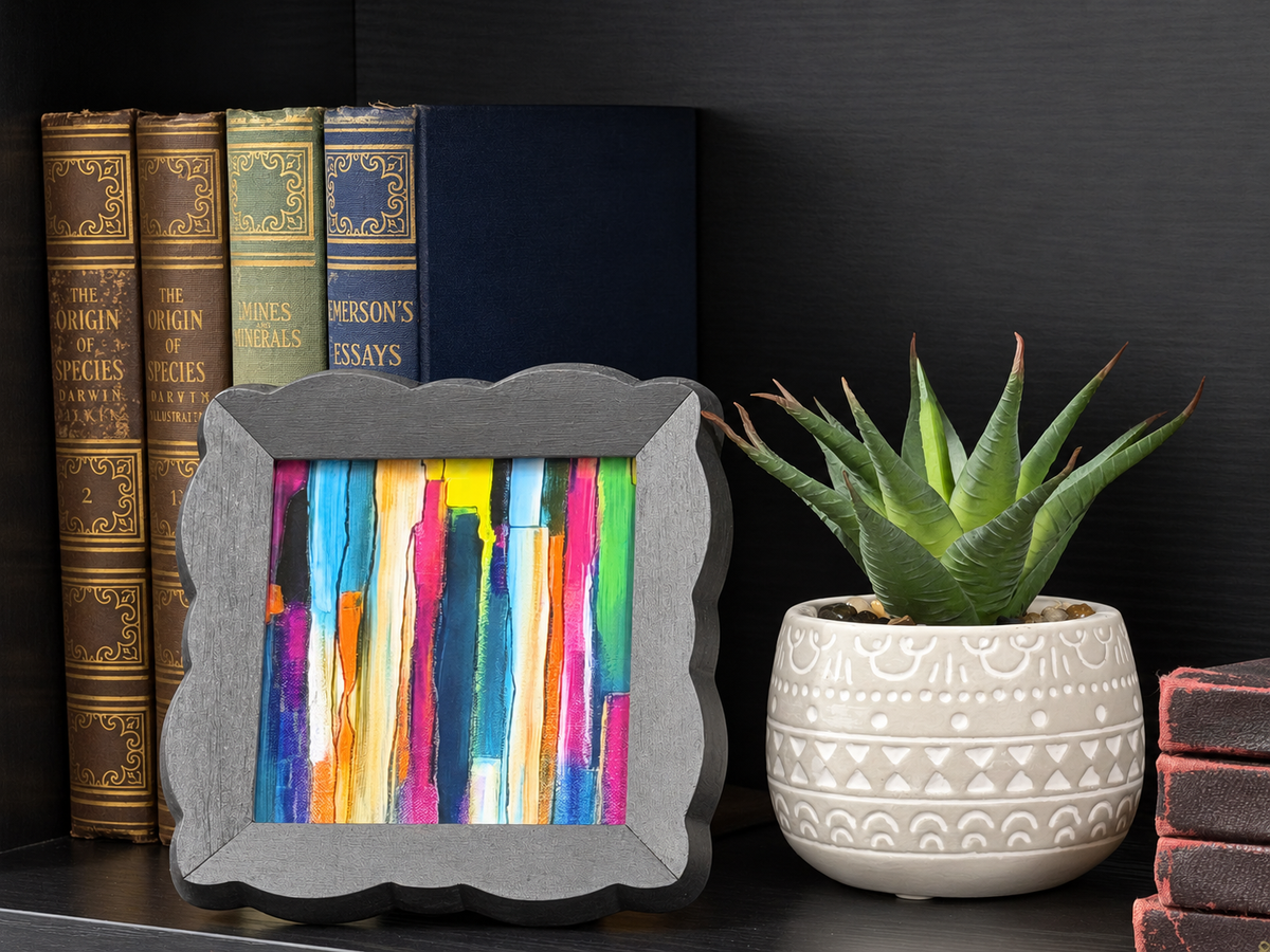 A square gray picture frame with scalloped edges holding a colorful abstract print resembling book spines, styled on a dark shelf next to a potted succulent and books.