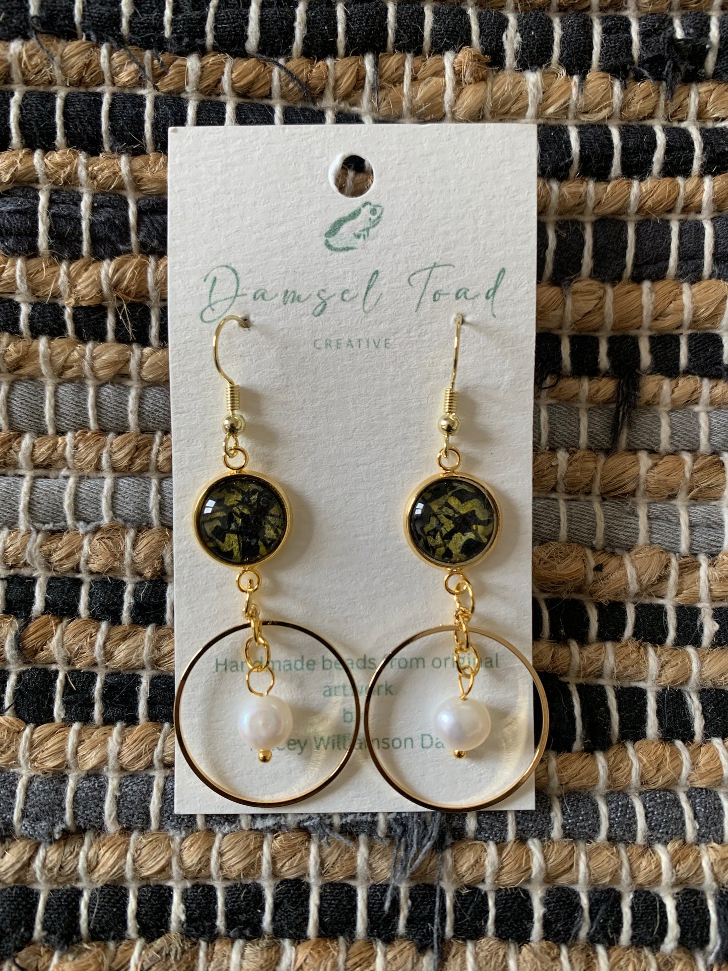 Pair of earrings on a 'Damsel Toad Creative' branded card against a woven fabric background.