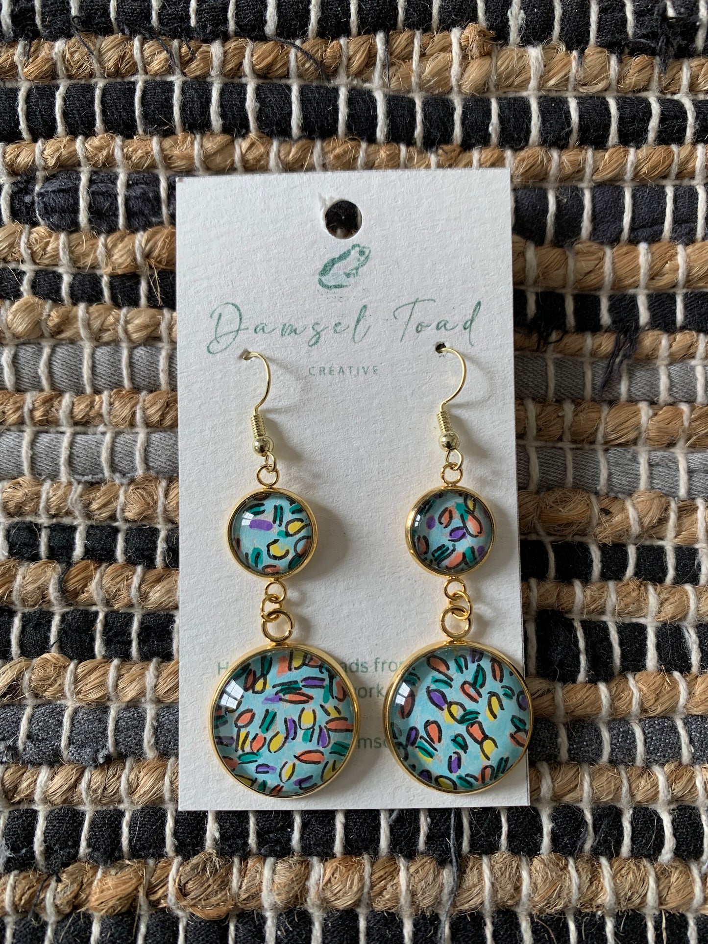 Pair of earrings on a 'Damsel Toad' branded card against a woven fabric background