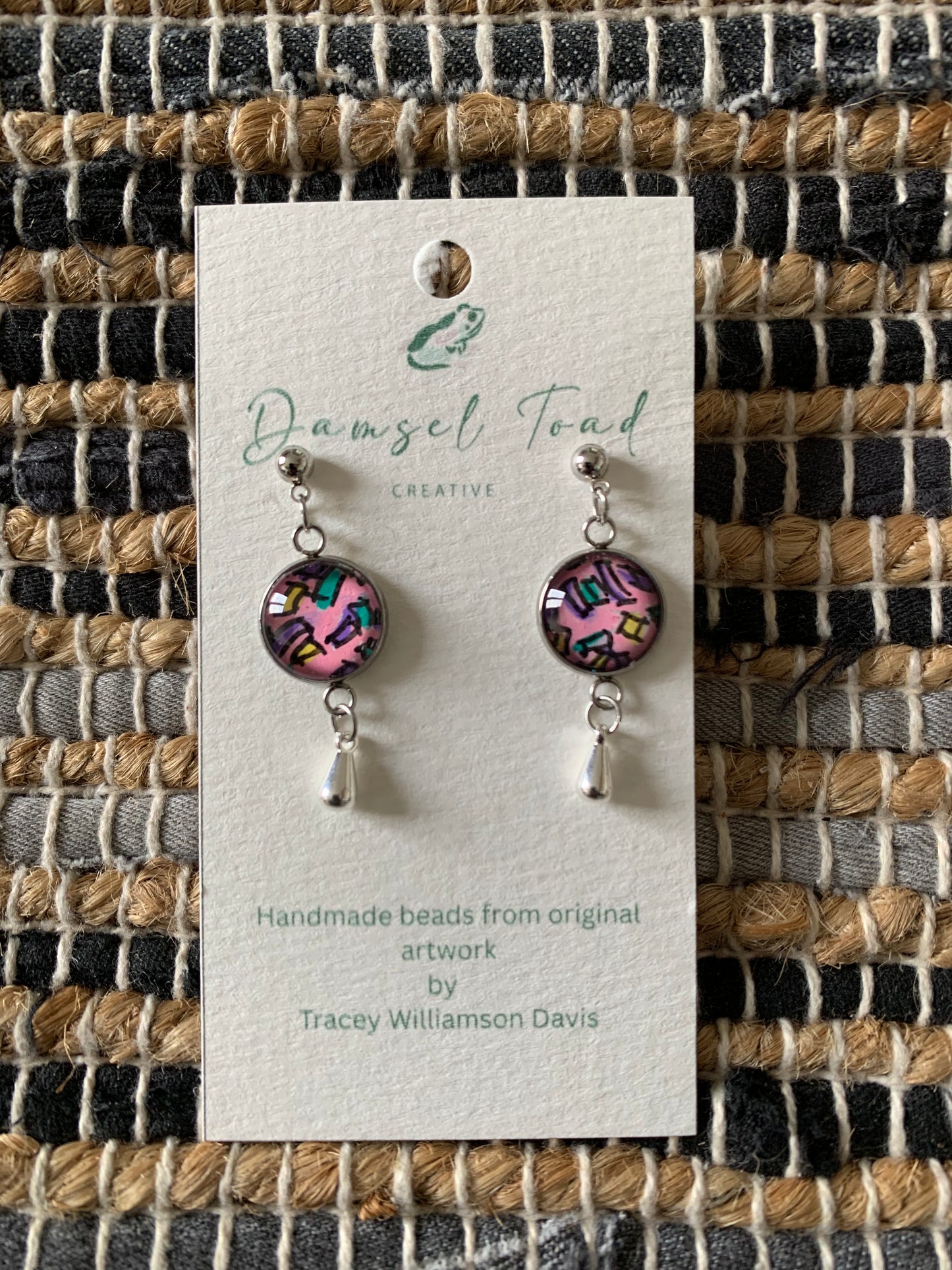 Pair of earrings on a 'Damsel Toad Creative' card against a woven fabric background