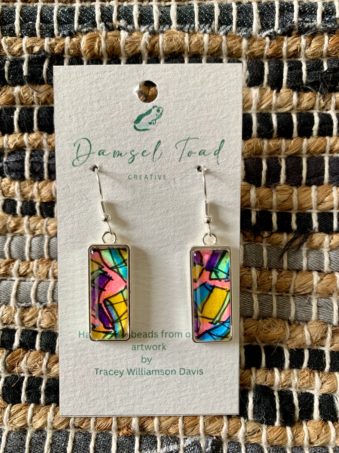 Colorful rectangular earrings on a Damsel Toad Creative card against a woven fabric background