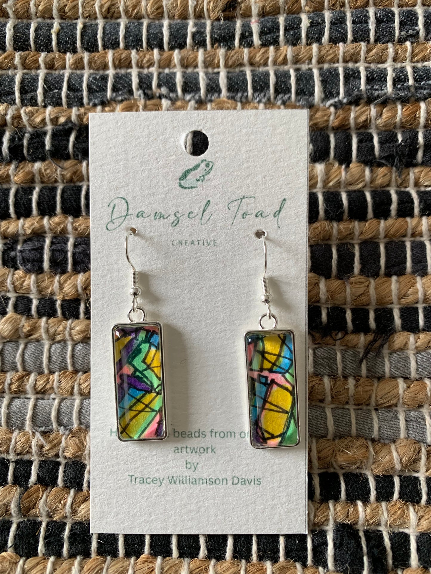 Colorful rectangular earrings on a Damsel Teal Creative card against a woven fabric background