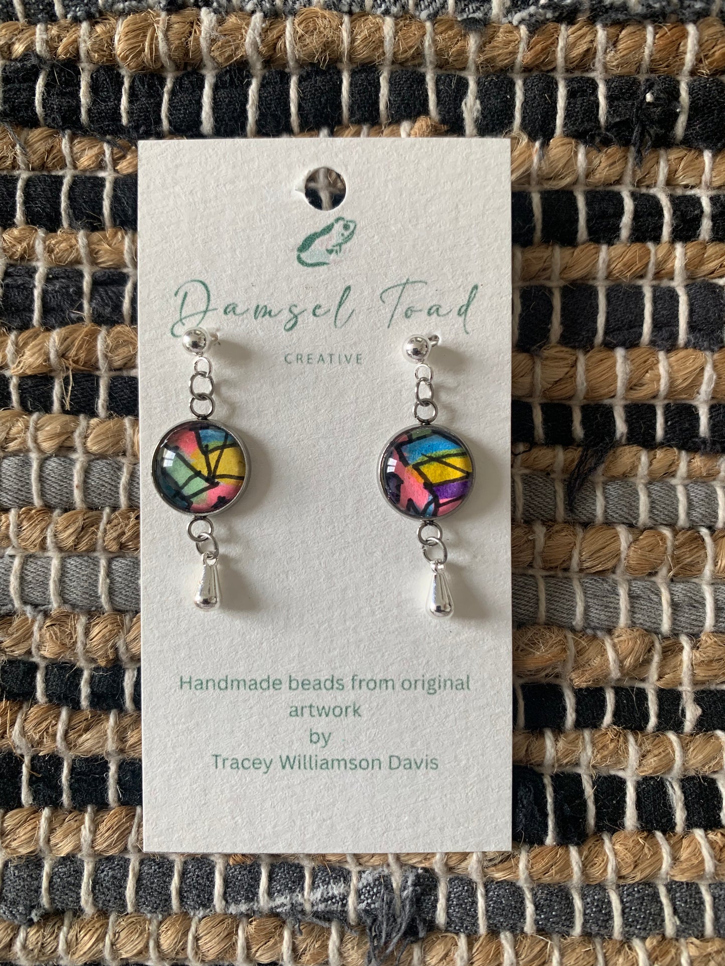Pair of colorful earrings on a Damsel Toad Creative card against a woven fabric background.