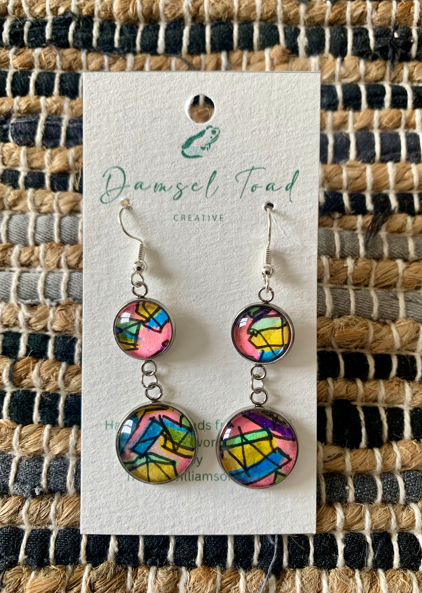 Colorful earrings on a 'Damsel Toad Creative' branded card against a woven fabric background.