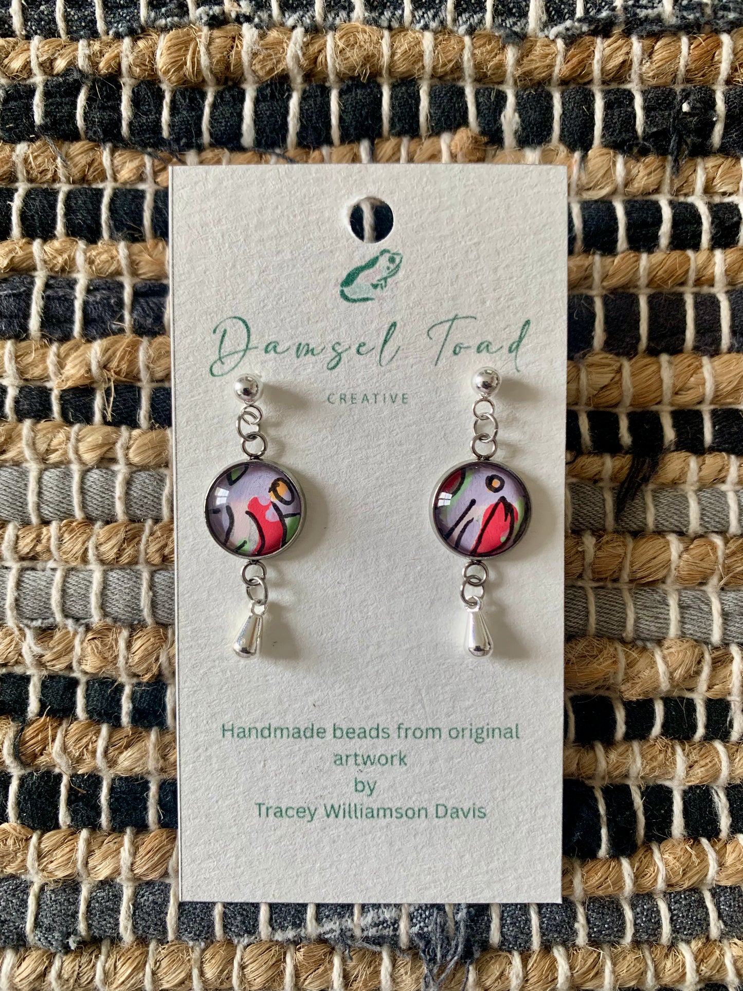 Pair of earrings on a Damsel Toad Creative card against a woven fabric background