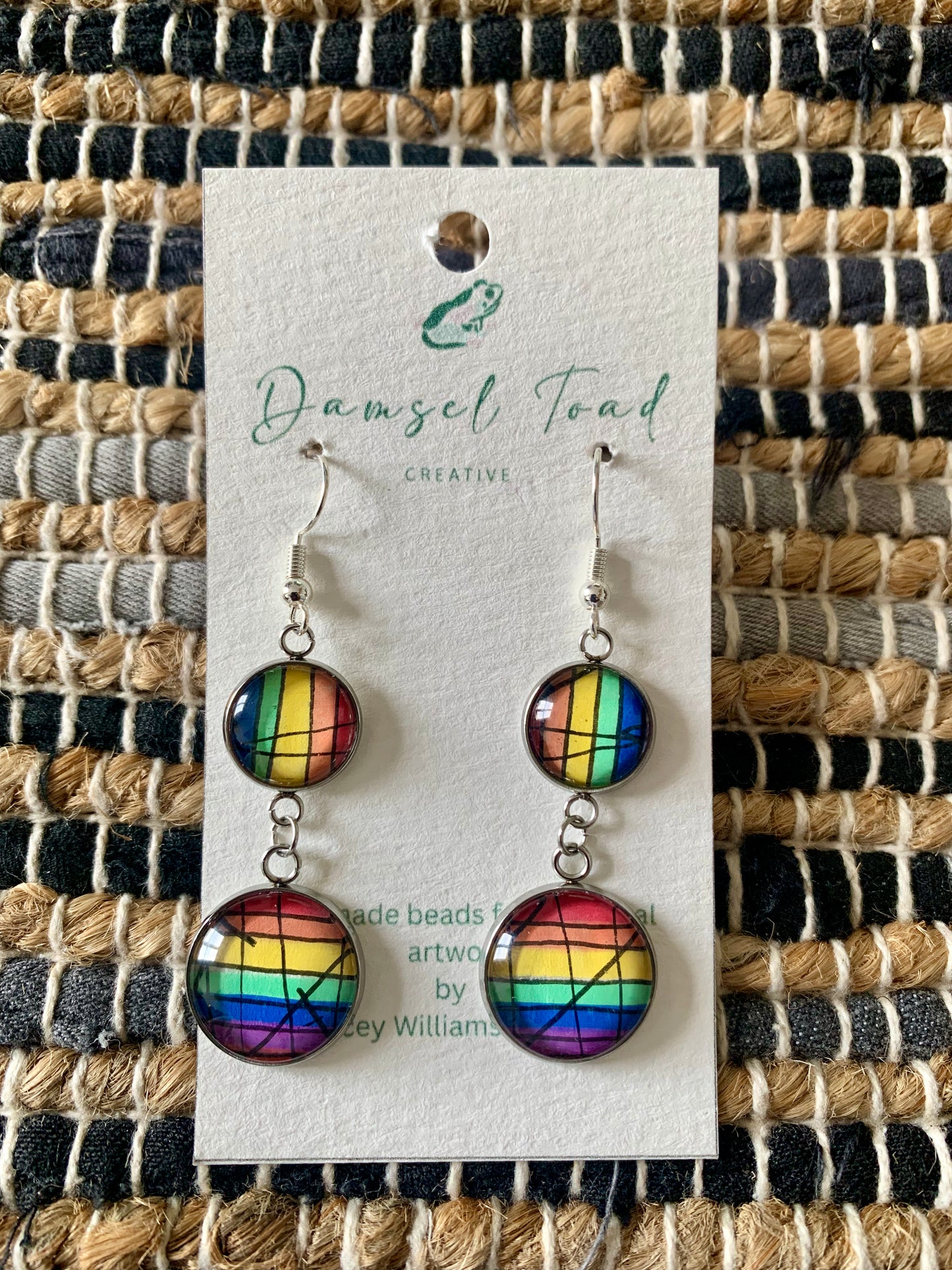 Rainbow patterned earrings on a Damsel Toad Creative card against a woven fabric background