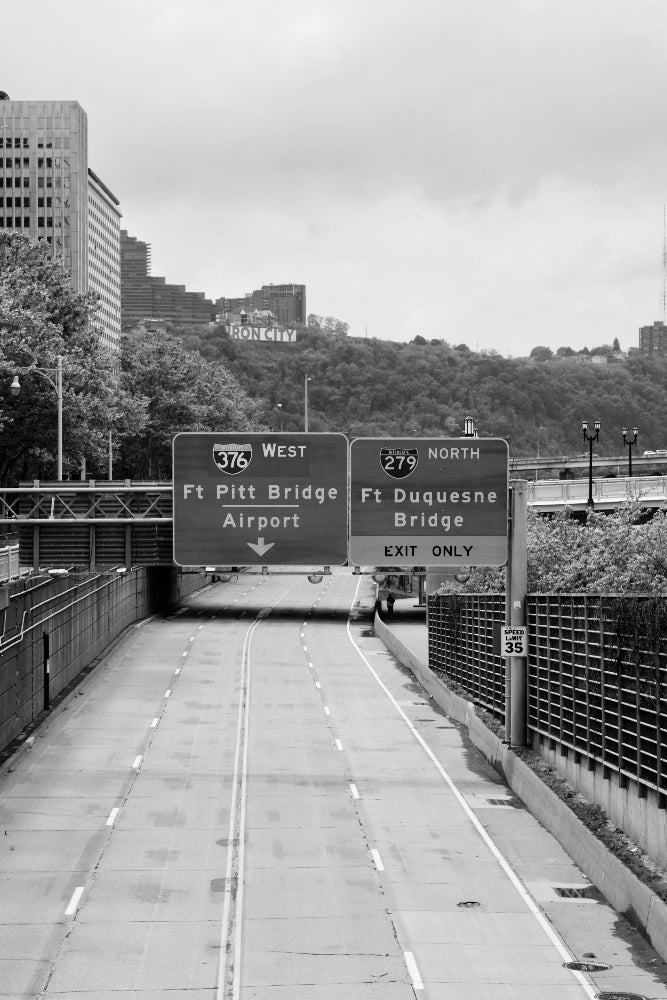 Highway with road signs indicating directions to bridges, surrounded by urban buildings and trees.