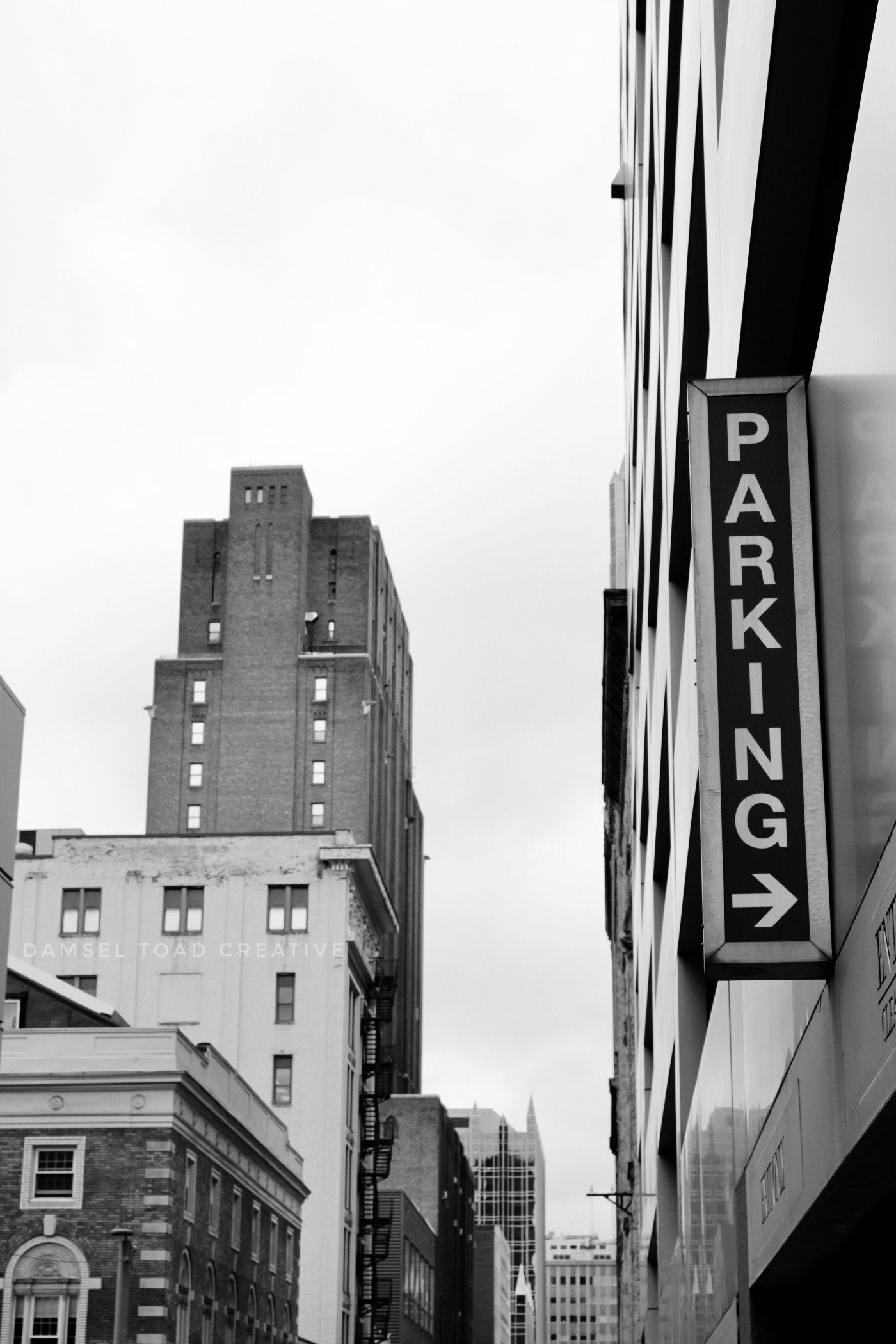 A black and white photo of a 'PARKING' sign on a city building's side, with a blurred background of other buildings.