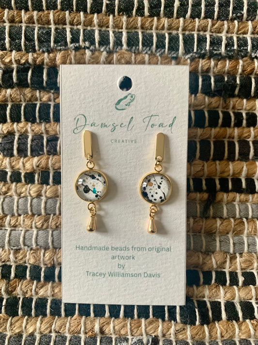Pair of gold earrings on a Damsel Toad Creative card against a woven fabric background