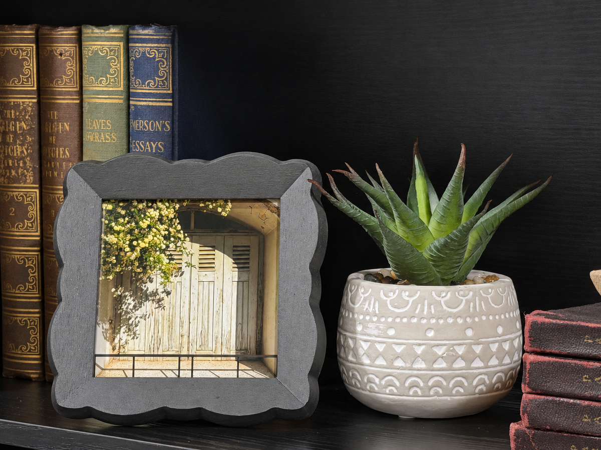 A square gray picture frame with a scalloped edge holding a photo of a wooden door, styled on a dark shelf next to a potted succulent and antique books.