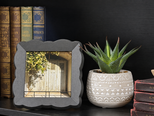 A square gray picture frame with a scalloped edge holding a photo of a wooden door, styled on a dark shelf next to a potted succulent and antique books.
