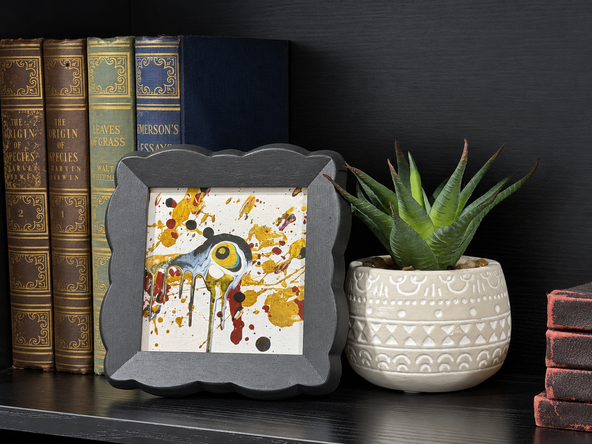 A square framed art print featuring a stylized bird with a large yellow eye surrounded by yellow, red, and black paint splatters on a white background, styled on a dark bookshelf next to a potted succulent.