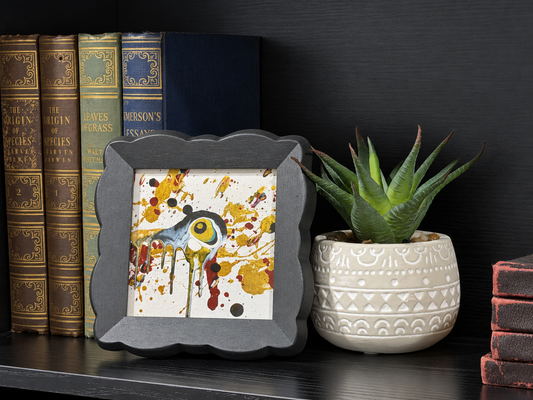 A square framed art print featuring a stylized bird with a large yellow eye surrounded by yellow, red, and black paint splatters on a white background, styled on a dark bookshelf next to a potted succulent.