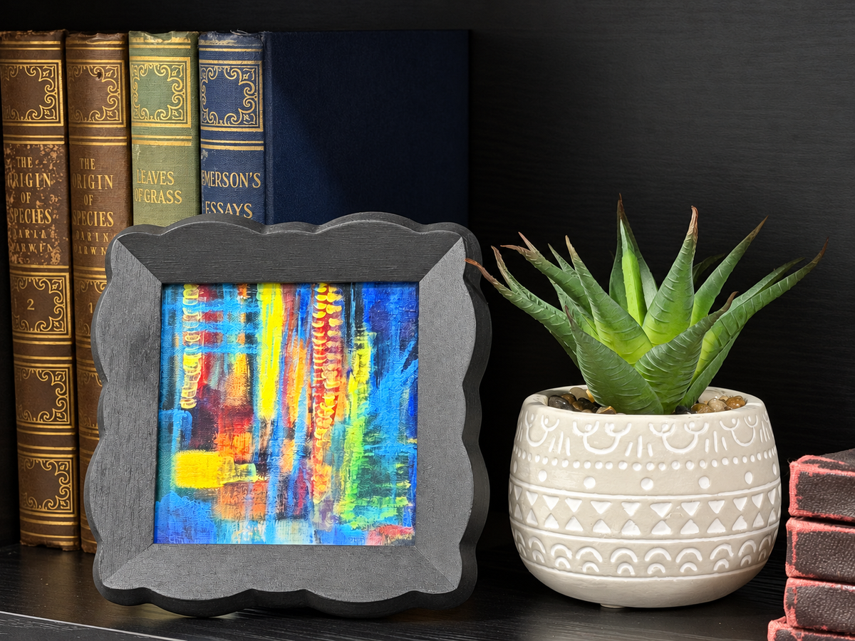 A square framed abstract painting with a dark gray scalloped frame, displayed on a shelf next to a potted succulent and books.