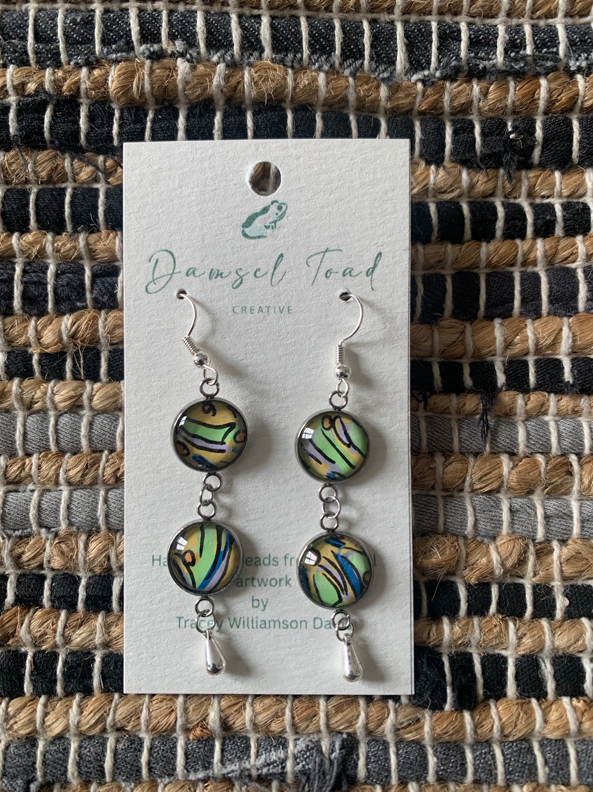 Pair of earrings on a 'Damsel Toad Creative' card against a woven fabric background