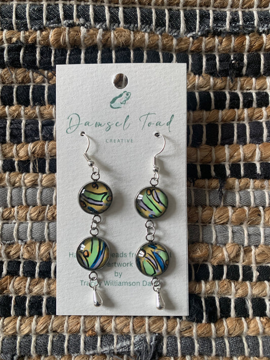 Pair of earrings on a 'Damsel Toad Creative' card against a woven fabric background