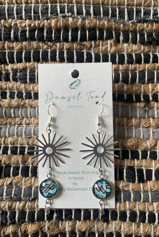 Sunburst-shaped earrings on a 'Damsel Toad Creative' card against a woven fabric background