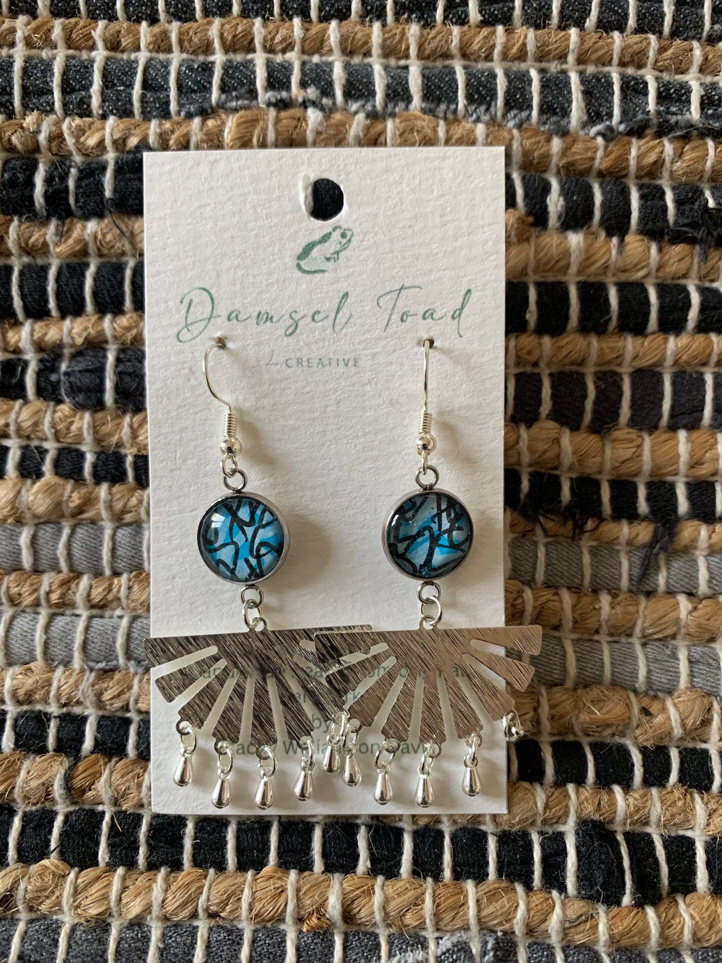 Pair of earrings on a 'Damsel Toad Creative' branded card against a woven fabric background.