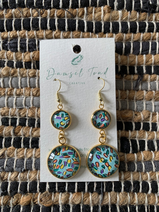 Pair of earrings on a 'Damsel Toad' branded card against a woven fabric background