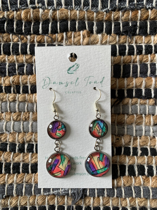 Colorful earrings on a 'Damsel Toad' branded card against a woven fabric background