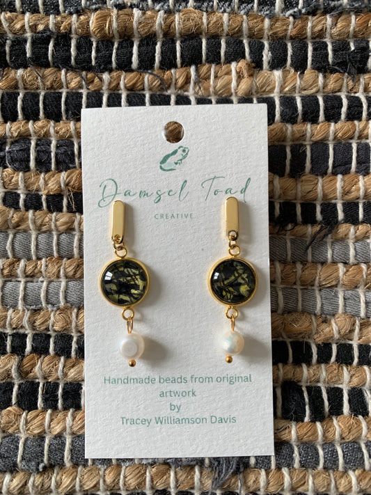 Gold earrings with black and white glass beads and pearl charms on a Damsel Toad Creative card against a woven fabric background.
