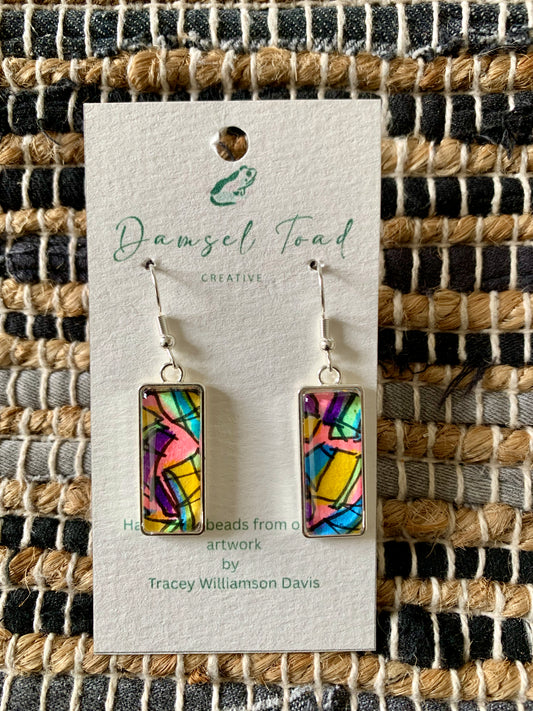 Colorful rectangular earrings on a Damsel Toad Creative card against a woven fabric background