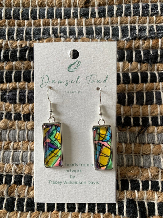 Colorful rectangular earrings on a Damsel Teal Creative card against a woven fabric background