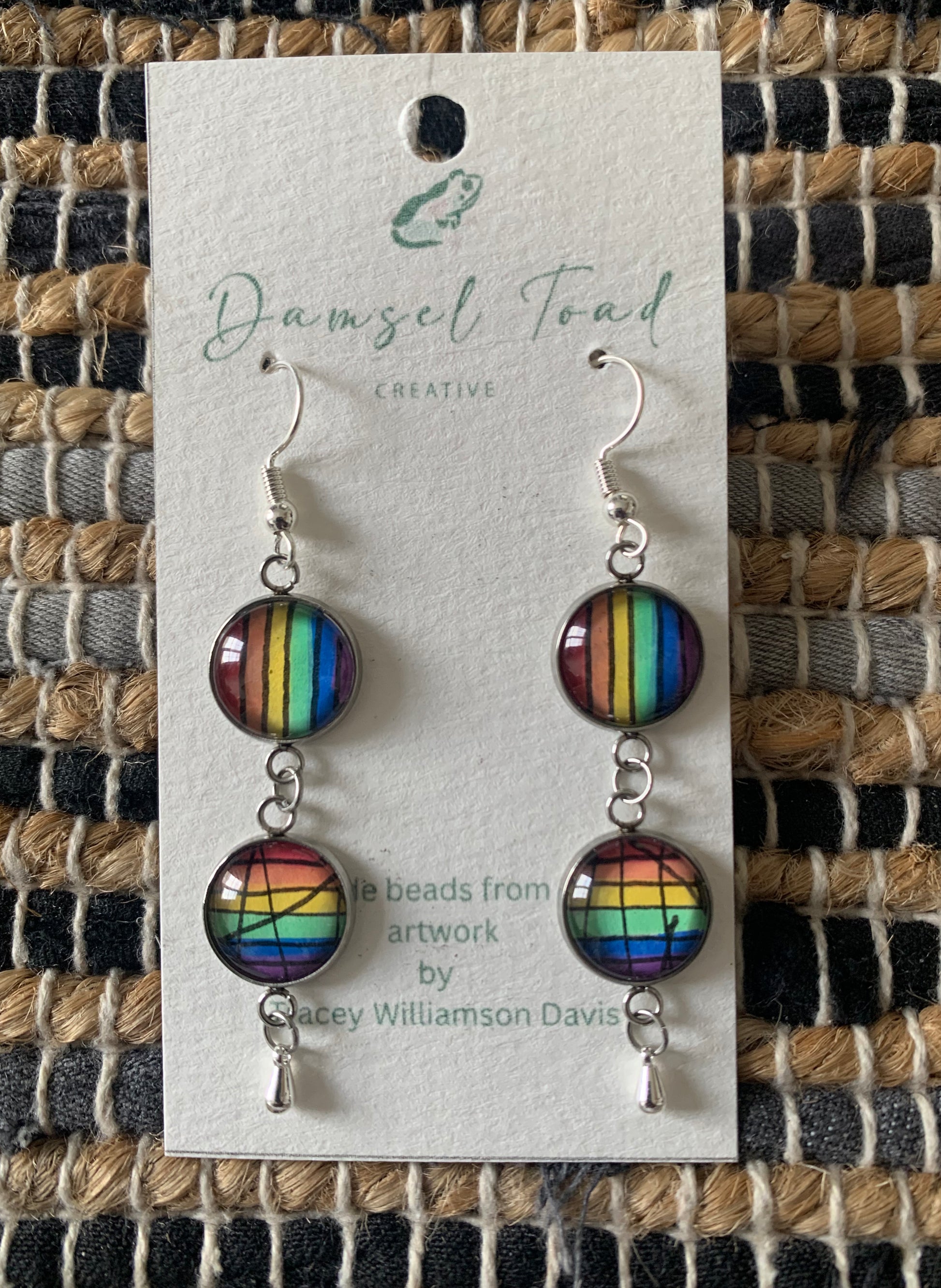 Rainbow striped earrings on a Damsel Toad Creative card against a textured fabric background