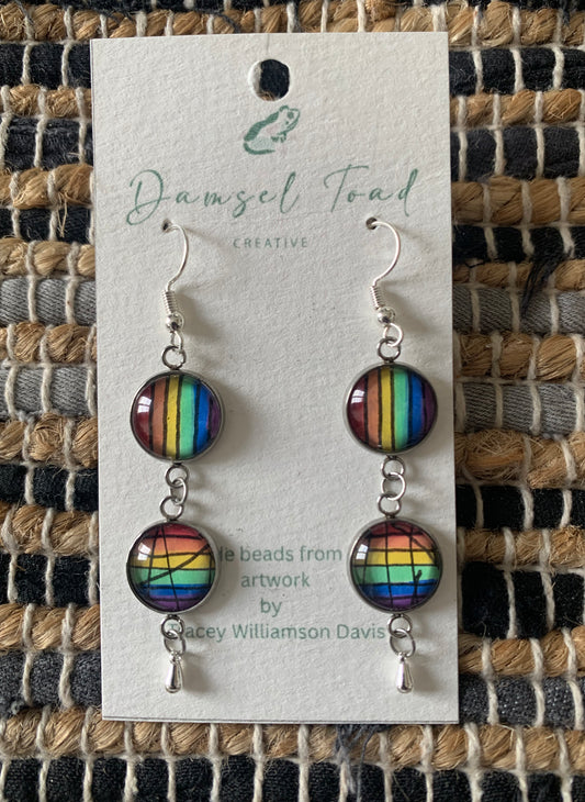 Rainbow striped earrings on a Damsel Toad Creative card against a textured fabric background