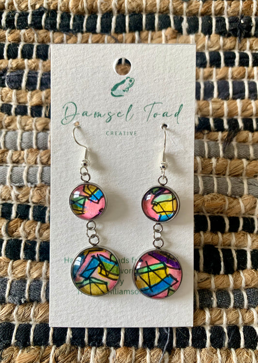 Colorful earrings on a 'Damsel Toad Creative' branded card against a woven fabric background.