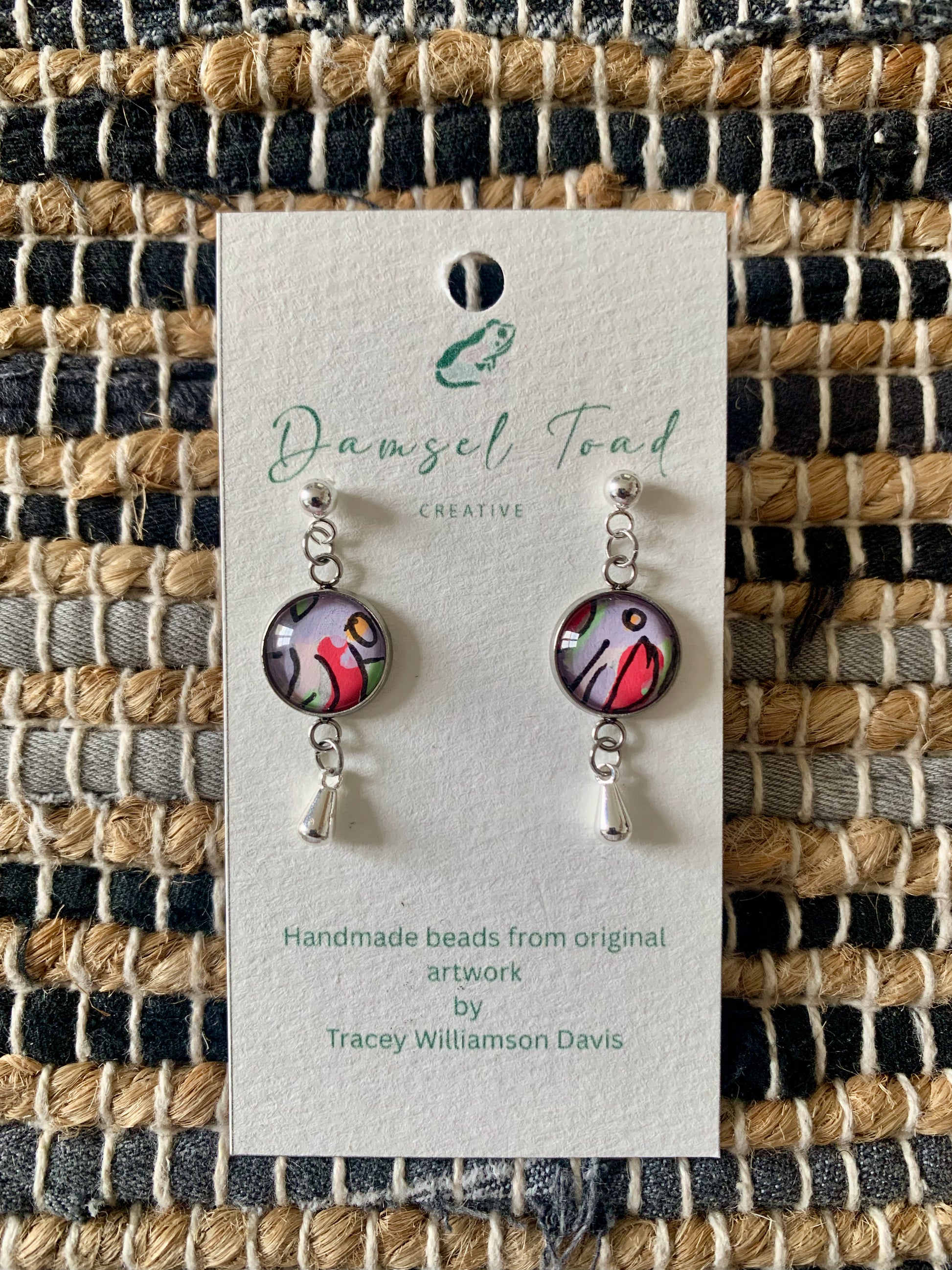 Pair of earrings on a Damsel Toad Creative card against a woven fabric background