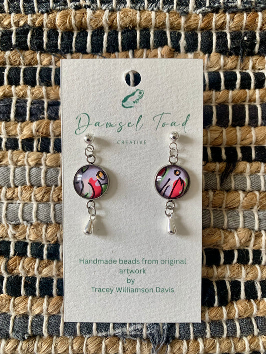 Pair of earrings on a Damsel Toad Creative card against a woven fabric background