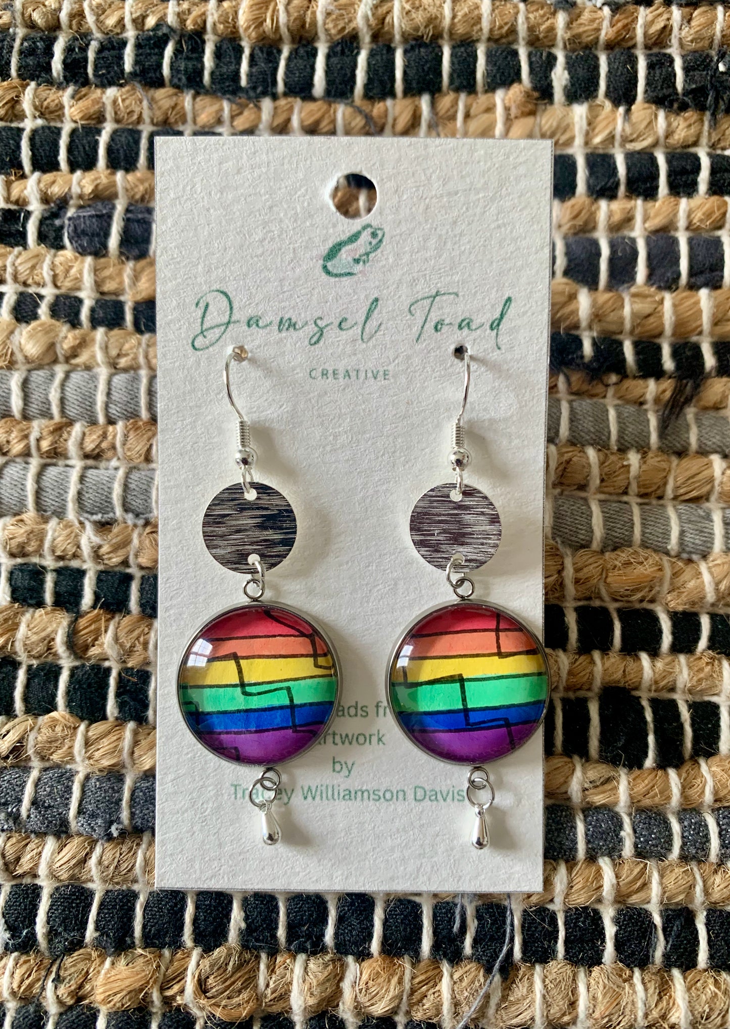 Rainbow earrings on a Damsel Toad Creative card against a woven mat background