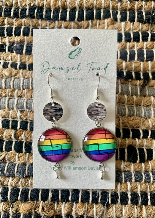 Rainbow earrings on a Damsel Toad Creative card against a woven mat background
