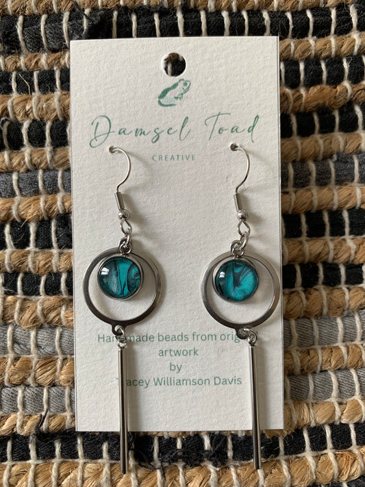 Pair of earrings on a 'Damsel Toad Creative' card against a woven fabric background