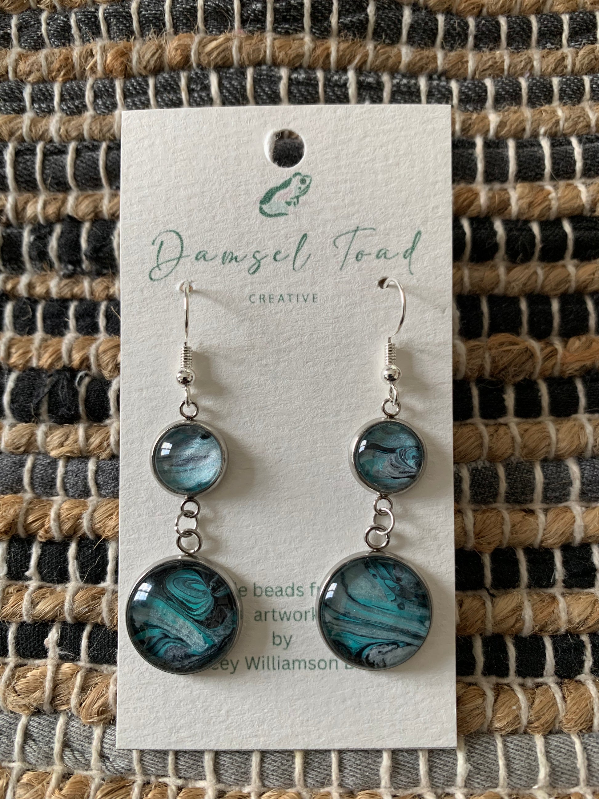 Pair of earrings on a 'Damsel Toad Creative' branded card against a textured fabric background.