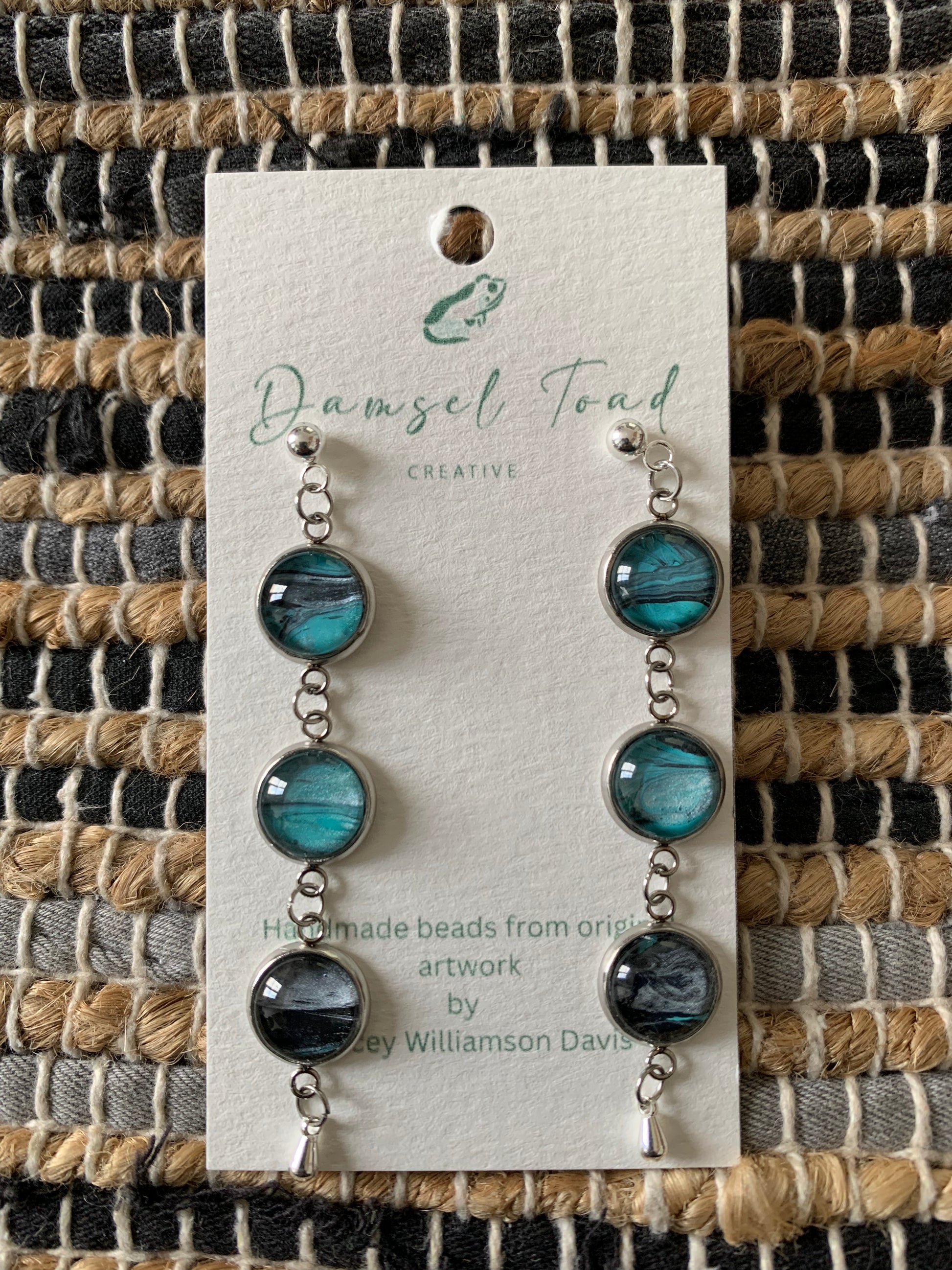 Pair of earrings on a Damsel Toad Creative card against a woven fabric background