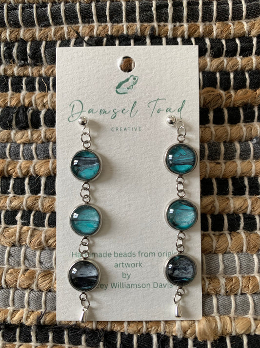 Pair of earrings on a Damsel Toad Creative card against a woven fabric background