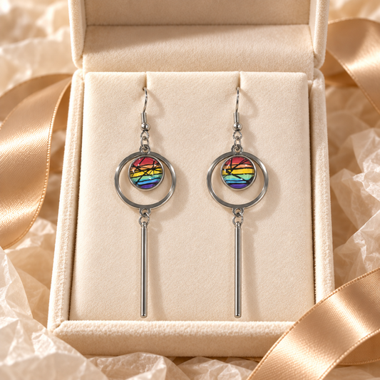 A pair of silver-toned drop earrings with circular hoops and vertical bars, each featuring a colorful circular inset with a rainbow sunset design, displayed inside an open cream jewelry box with satin ribbon.