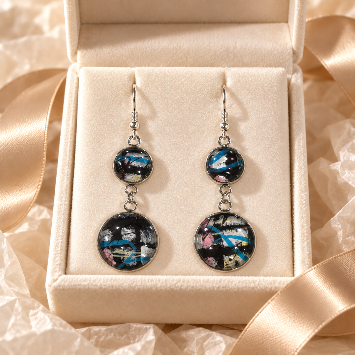 A pair of double-drop statement earrings with circular black and blue marbled resin pendants, displayed in a cream-colored jewelry box with satin ribbon.