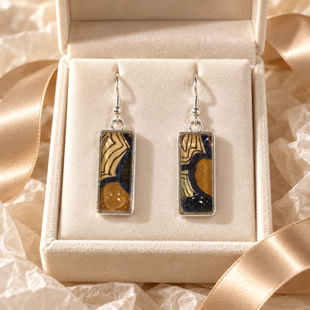 A pair of silver-tone rectangular drop earrings with abstract paintings under glass in black, brown, and cream, presented in an open cream jewelry box with satin ribbon.