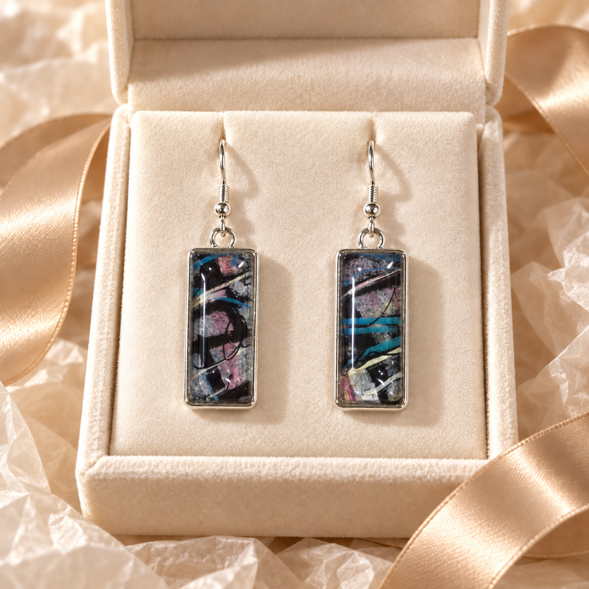 A pair of rectangular drop earrings with silver-toned metal frames and hooks, featuring an abstract glass design in black, blue, pink, and white. The earrings are displayed inside an open beige velvet jewelry box with a champagne satin ribbon.