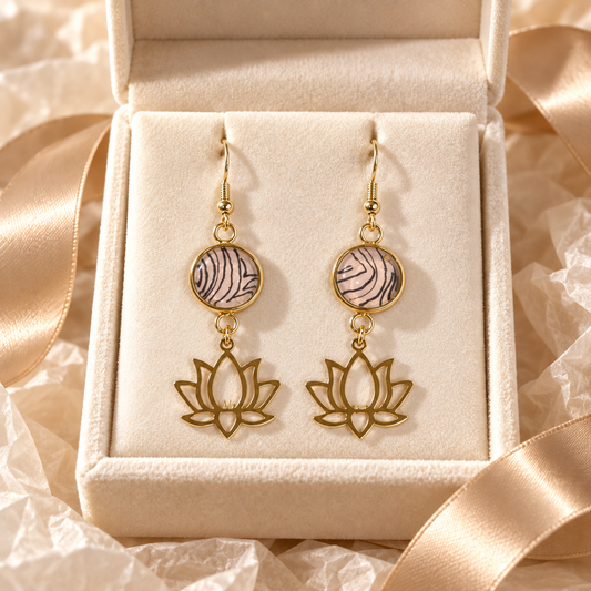 A pair of gold-tone dangle earrings displayed in an open cream jewelry box. Each earring has a round top section with a pink and gray zebra-stripe pattern, hanging from a gold hook, and a lower lotus flower outline charm.