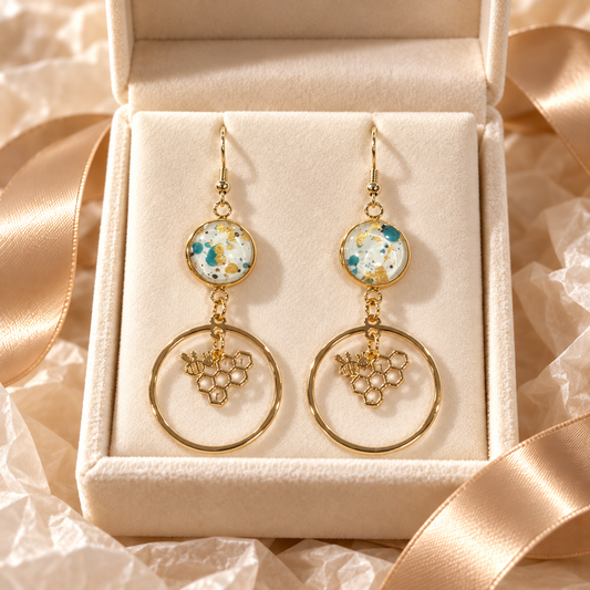 A pair of gold dangle earrings displayed in an open cream jewelry box with satin ribbon. Each earring has a round glass bead top section with a  blue, white, and gold pattern, a small gold bee and hive charm inside a large circular gold hoop dangling below.